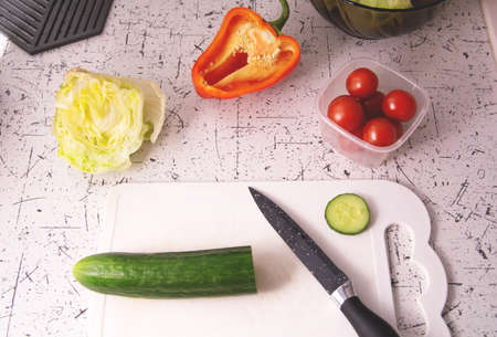 Ingredients for preparing salad on the kitchen table. Salad bowl with lettuce, red tomatoes, green cucumbers and lettuce. Knife on a cutting boardの写真素材