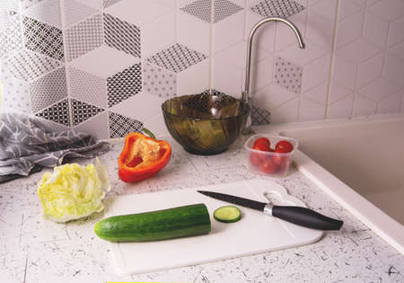Ingredients for preparing salad on the kitchen table. Salad bowl with lettuce, red tomatoes, green cucumbers and lettuce. Knife on a cutting boardの写真素材