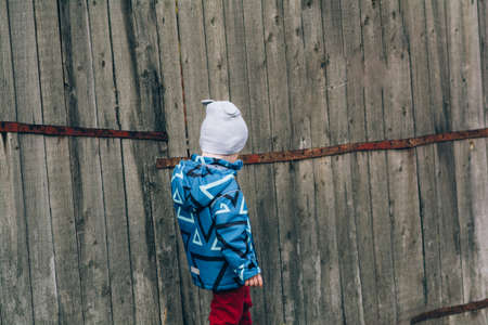 A child in a blue jacket and a hat walks near a wooden wall. Real and serene people, calmness and relaxation. Capturing the momentの写真素材