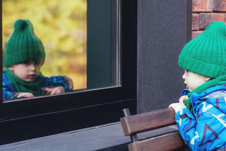A small child looks out the window from the street. The boy sees his reflection in the mirror windowの写真素材
