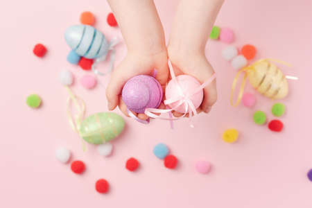 Children's hands hold Easter shiny eggs. Decorative eggs and fluffy balls on a pink background. Background for your project for the holiday happy easterの写真素材