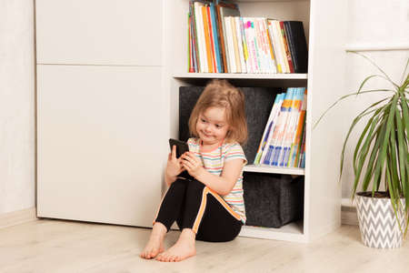 Little child sits on the floor in his room and uses the phone. Smiling girl looking at the phone. Childhood communication, technology and communication conceptの写真素材
