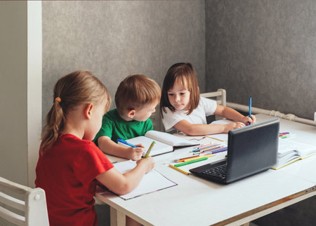 Children sit at a table with pens and notebooks and use a laptopの写真素材