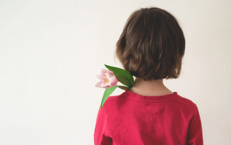 Child from the back holds a pink tulip on his shoulder on a white background. Girl with short hair in a red t-shirt. copy spaceの写真素材
