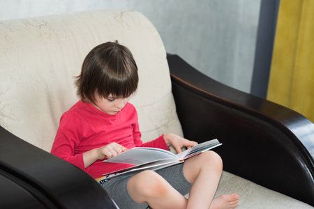 Cute baby sitting on the armchair and studying a book Leisure and education at home. child portrait looking into a bookの写真素材