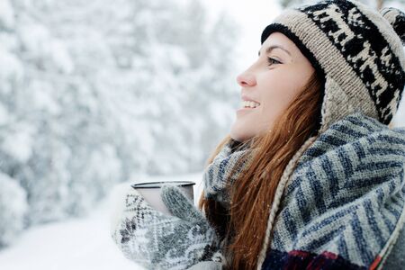 Beautiful young woman standing among snowy trees in winter forest and enjoying cup of hot tea.
