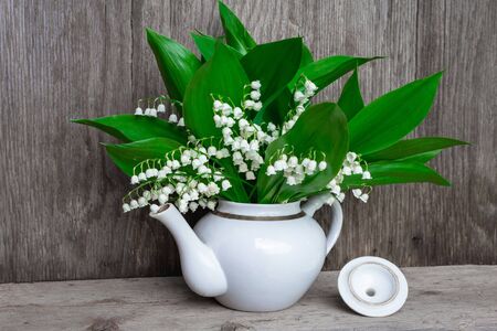 lilies of the valley stand in a white teapot on a wooden background, front view