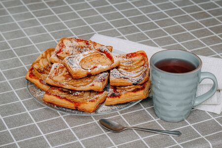 puffs of puff pastry with Apple, cinnamon and powdered sugar, in a plate on the table with a checkered tablecloth, red tea in a turquoise mug, a spoonの写真素材