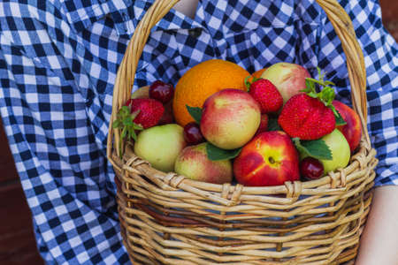 the girl's hands hold nectarines, oranges, strawberries, cherries and mint leaves, which lie in a wicker basket. wooden backgroundの写真素材