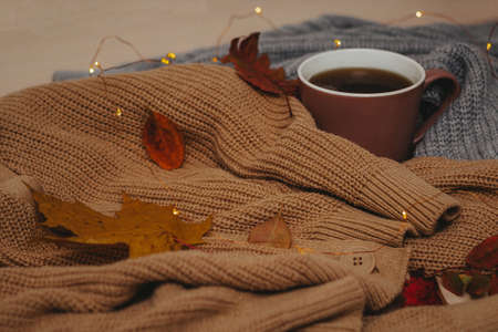 on the table are two cozy sweaters rolled up around a hot cup of coffee, autumn dry leaves are...