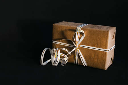 Gift box wrapped in recycled craft paper and tied with a white and gold ribbon bow on a black...