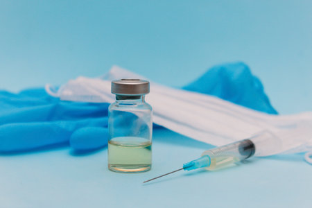 The vaccine is in a jar next to a syringe, a medical mask and blue gloves on a blue background.