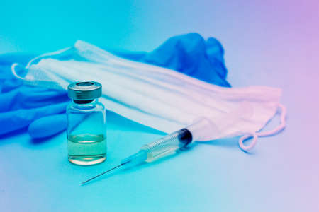 The vaccine is in a jar next to a syringe, a medical mask and blue gloves on a blue background.の写真素材