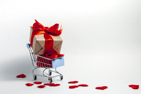 A supermarket cart filled to the brim with red confetti hearts and a gift box wrapped in kraft paper and tied with a red ribbon on a white background. The concept of buying presents. Copy space.の写真素材