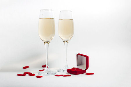 Two glasses of champagne and a red box ring on a white minimalist background with red confetti hearts. copy space.の写真素材