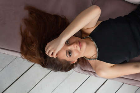A happy, beautiful, successful woman with long hair lay down on a sofa in a black dress. Portrait,...