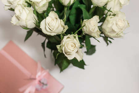 White tea roses stand in a glass vase, which is tied with a red ribbon and stands on the table. There is a gift nearby. in a pink box. Gift bouquet.の写真素材