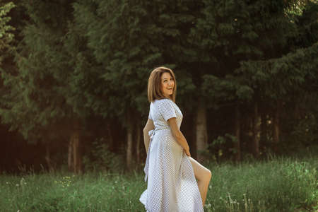 A happy young woman with short hair in a white polka dot dress walks in a meadow near the forest. Portrait of a girl with natural beauty. Selective Focus.の写真素材