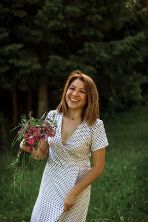 A beautiful happy girl in a white dress holds a bouquet of wild flowers in her hands and smiles.Portrait of a cheerful girl with a bouquet of wild flowers in a meadow near the forest. selective focus.の写真素材
