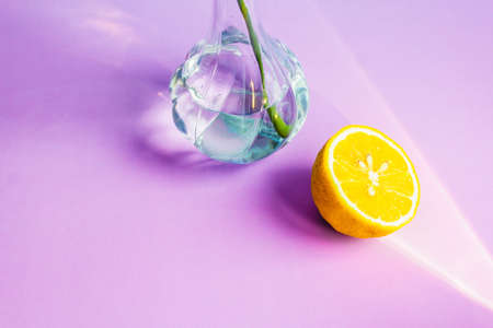 Half of a yellow lemon lies next to a glass vase with water on a lilac background of waters by the...