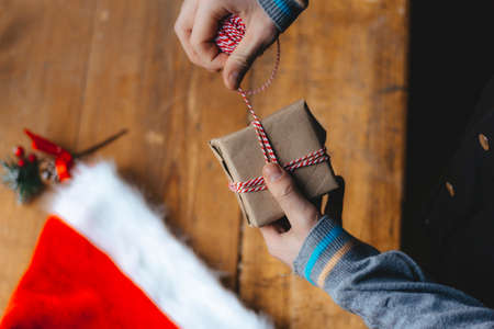 Christmas gift packaging. Hands wrap a small gift box in craft paper and a red-white string on the...