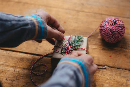 Christmas gift packaging. Men's hands in a blue sweater are packing a small gift box on a wooden table. Top view. Selective focus.の写真素材
