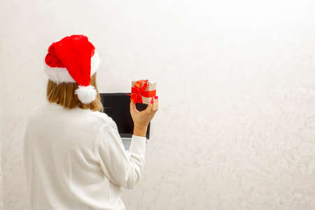 A girl in a Santa hat stands with her back against a light background and holds a laptop and a Christmas gift with a red bow in her hands. Christmas online shopping of goods and gifts.の写真素材