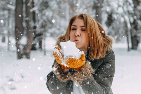 A happy young girl blows on the white snow, which lies on cozy knitted woolen mustard-colored...