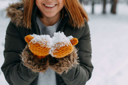 A happy young girl holds white snow on knitted mustard-colored woolen mittens in a snowy forest. Christmas holidays, Winter time. without a face.の写真素材
