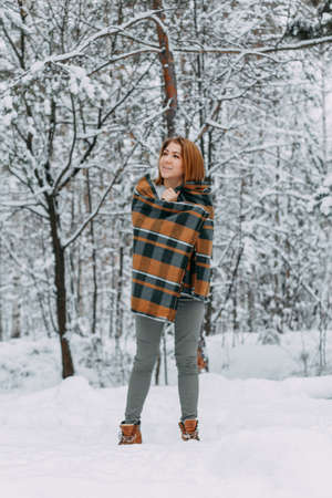 A happy young girl, wrapped in a plaid plaid and basking on a walk in the winter forest. A walk in...