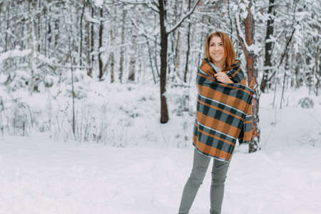 A happy young girl, wrapped in a plaid plaid and basking on a walk in the winter forest. A walk in...