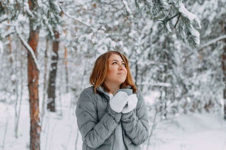 A beautiful girl in a gray jacket and knitted white mittens drinks a drink in a snowy forest, looks up and dreams. Comfort and warmth in the winter season.の写真素材