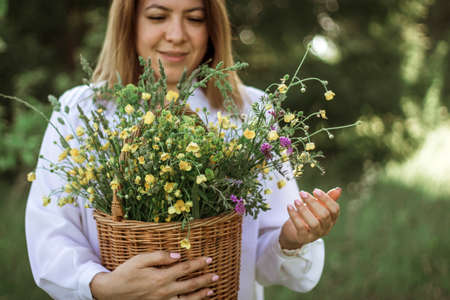 A girl in a white blouse holds a wicker basket with a bouquet of wild flowers. Summer walk in the field. midsection.の写真素材
