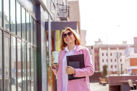 a young beautiful brunette businesswoman in sunglasses and a pink coat is walking along a city...
