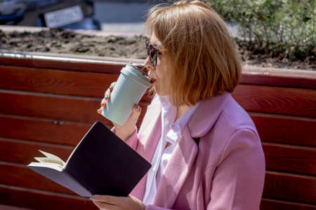 A young beautiful girl in sunglasses drinks coffee from an eco-friendly thermocup and holds a...