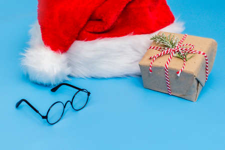 A Santa Claus hat, a Christmas gift tied with string, and round glasses on a blue background. A...