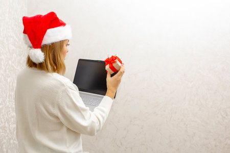 A girl in a Santa hat stands with her back against a light background and holds a laptop and a...