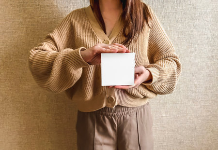 An unrecognizable brunette girl in a beige sweater holds a white square mockup in her hands. An empty sign in the hands of a girl.の写真素材