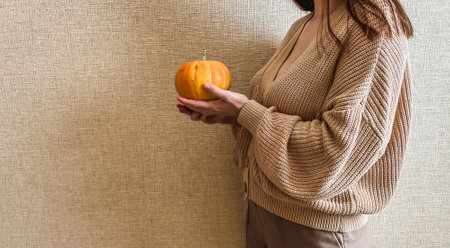 An unrecognizable brunette girl in a beige sweater holds a bright orange pumpkin in her hands on a beige background.の写真素材