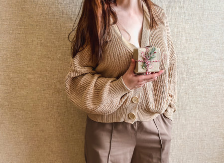 An unrecognizable brunette girl in a beige sweater holds a Christmas gift in eco packaging in her hands. a small gift in honor of Christmas, new year.の写真素材