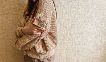 An unrecognizable brunette girl in a beige sweater holds a Christmas gift in eco packaging in her hands. a small gift in honor of Christmas, new year.の写真素材