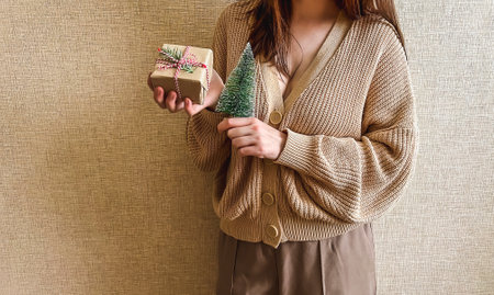 An unrecognizable brunette girl in a beige sweater holds a Christmas gift in eco packaging and a green Christmas tree in her hands. a small gift in honor of Christmas, new year.の写真素材