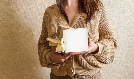 An unrecognizable brunette girl in a beige sweater holds a white square mockup and a gift in her...
