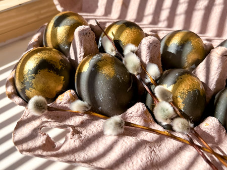 Stripes of light and shadow from the blinds lie on the black and gold Easter eggs that are in the...