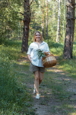 A young beautiful woman walks along a path in the forest with a wicker basket and a bouquet of flowers and herbs.の写真素材