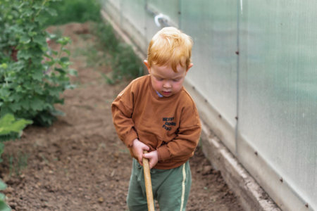 A little red-haired boy holds a rake in the garden, digging the ground. Summer in the village.の写真素材
