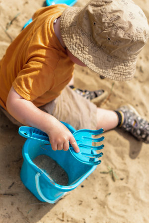 0A little boy in a red T-shirt and a sand hat with a blue bucket is sitting on the sand and playing.の写真素材