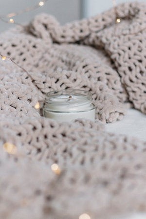 A candle in a glass jar with a wooden wick stands among a knitted plush soft warm plaid. Warm...
