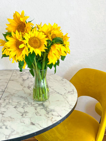 A bouquet of sunflowers stands in a glass vase on a marble table in a white kitchen with yellow armchairs. The kitchen interior is decorated in white tones.の写真素材