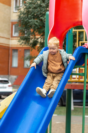 A little boy in a sweatshirt and jacket slides down a bright blue slide on a playground in the city. A walk in the fresh air, sincere emotions. Vertical photo.の写真素材